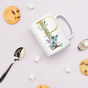 Può includere: Tazza in ceramica bianca con una lettera P dorata e un design floreale blu acquerellato. La tazza è su una superficie bianca con cubetti di zucchero e biscotti.