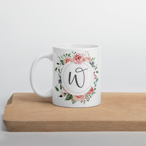May include: White ceramic mug featuring a floral design with the letter "W" in a circle. The flowers are pink and green.