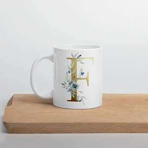 Può includere: Tazza in ceramica bianca con una lettera F dorata circondata da fiori ad acquerello blu e verdi.