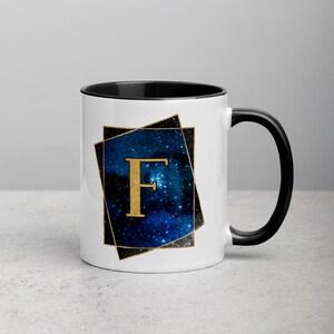 Può includere: Tazza in ceramica bianca con bordo e manico neri. La tazza presenta un design a tema galattico con una lettera "F" dorata al centro.