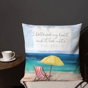 May include: A throw pillow with a watercolor beach scene. The pillow features a yellow umbrella, a red and white striped beach chair, and the text "I followed my heart and it led me to THE BEACH".
