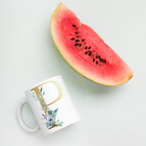 Può includere: Una tazza in ceramica bianca con una lettera dorata "P" e un design floreale blu acquerellato. La tazza è accanto a una fetta di anguria.