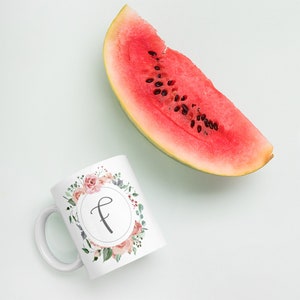 Può includere: Una tazza in ceramica bianca con un design floreale e la lettera "F" al centro. La tazza è su una superficie bianca accanto a una fetta di anguria.