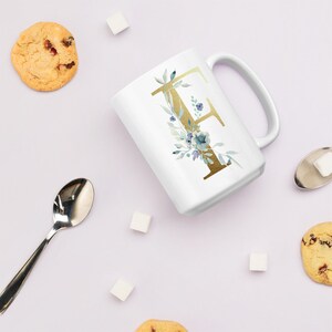 Può includere: Tazza in ceramica bianca con una lettera dorata "F" e un design floreale ad acquerello. La tazza è su una superficie bianca con cubetti di zucchero e biscotti.