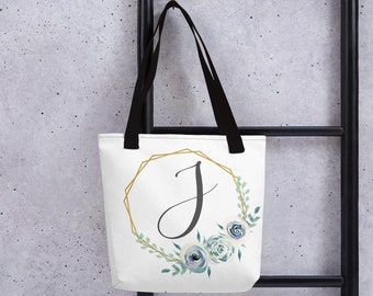 May include: White tote bag with black handles featuring a gold geometric frame with the letter "J" in black script. The frame is adorned with blue and green watercolour floral accents.