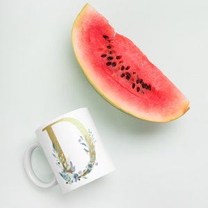 May include: White ceramic mug with a gold letter D surrounded by blue and green watercolor floral design. A slice of watermelon is in the background.