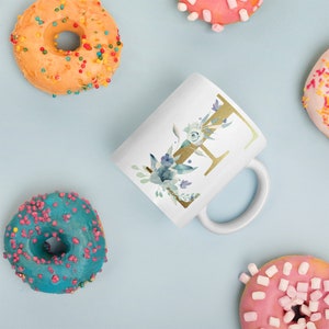 Könnte beinhalten: Weiße Keramiktasse mit einem goldenen Buchstaben "E" und einem Aquarell-Blumenmuster. Die Tasse ist von Donuts und anderen Dekoartikeln umgeben.