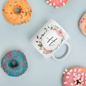 Puede incluir: Taza de cerámica blanca con un diseño floral y la letra "H" en un círculo. La taza está rodeada de donuts con chispas de colores.