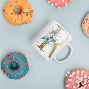 May include: White ceramic mug featuring a gold letter "R" and a blue watercolour floral design. The mug is surrounded by donuts with colourful sprinkles.