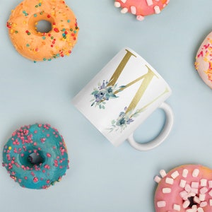 May include: White ceramic mug featuring a gold letter W and a blue watercolour floral design. The mug is surrounded by donuts with colourful sprinkles.