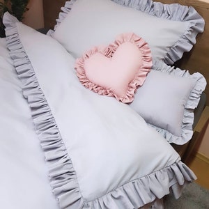 Könnte beinhalten: Ein grauer, gerüschter Bettbezug mit einem rosa herzförmigen Kissen und einem grauen, gerüschten Kissen darauf.