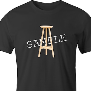 May include: A black t-shirt with the word "SAMPLE" printed in white across the front. A wooden stool is centered below the text.