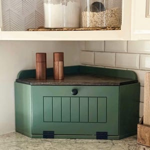 May include: A green corner bread box with a wooden top and a black knob. Two wooden salt and pepper grinders sit on top. Jars of flour and oats are on a shelf above the bread box. The kitchen has white subway tile and a patterned backsplash.