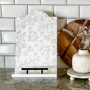 May include: A white wooden cookbook stand with a floral design, a wooden cutting board, and a small white vase with dried flowers. The items are arranged on a granite countertop in a kitchen setting.