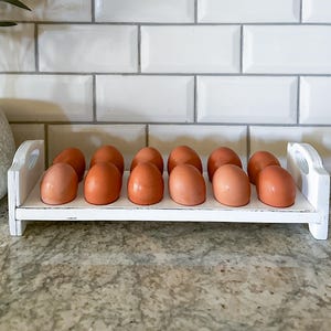 May include: White wooden egg holder with 12 brown eggs. The holder has two handles on the sides and is sitting on a countertop.
