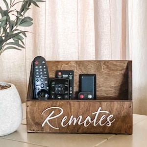 May include: A wooden remote control organizer with the word "Remotes" written on the front. The organizer is filled with four black remote controls.