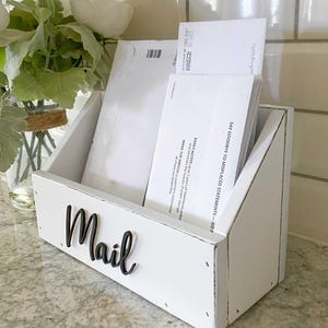 May include: White wooden mail organizer with the word "Mail" in black lettering. The organizer has three slots for mail and is sitting on a countertop.