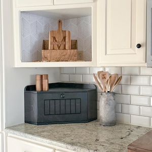 May include: A kitchen scene featuring a wooden cutting board with the name "Montgomery" engraved on it, displayed in a cabinet. A black bread box and a collection of wooden spoons in a gray vase are also visible.