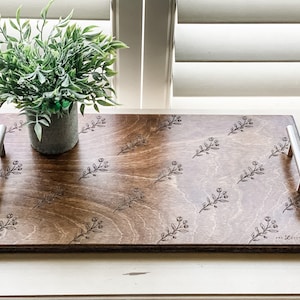 May include: A wooden serving tray with silver handles features a floral design engraved into the surface. The tray is brown and has a light wood grain pattern. The floral design is a repeating pattern of small branches with leaves and flowers.