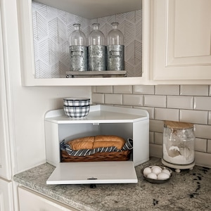 May include: A white corner bread box with an open door, revealing a loaf of bread in a wicker basket. Above, three glass bottles with metal tops sit on a shelf. A bowl and a jar with a wooden lid are also visible.