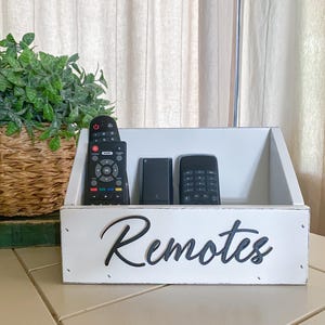 May include: White wooden box with the word "Remotes" written in black on the front. The box is holding four black remotes.