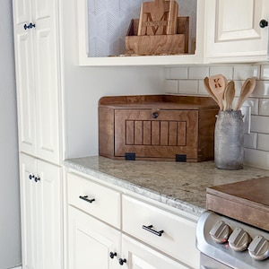 May include: A wooden corner bread box with a hinged door and black hardware sits on a granite countertop. Next to it, a gray textured crock holds wooden spoons, one marked with the letter K. A wooden cutting board labeled MONTGOMERY sits above.