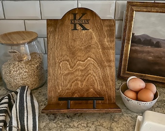 May include: A wooden cookbook stand with a black metal holder. The stand is stained a dark brown color and has the name "Krishan" engraved on the front.