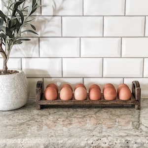 May include: A wooden egg holder with 12 brown eggs on a white tiled countertop. The holder is made of dark wood and has a rustic look.