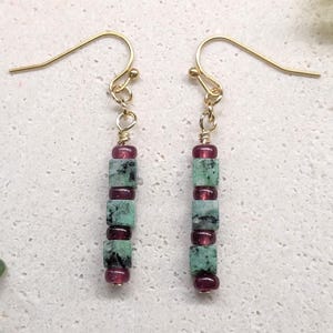 May include: A pair of dangle earrings featuring gold-tone metal hooks and wire. Each earring consists of a vertical arrangement of three square-shaped, mottled green stone beads alternating with three small, round, deep red translucent beads.
