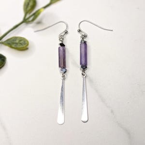 May include: A pair of dangle earrings featuring a silver hook, a cylindrical purple bead, and a silver teardrop-shaped charm. The earrings also have small black and gray beads. The earrings are displayed on a white surface.