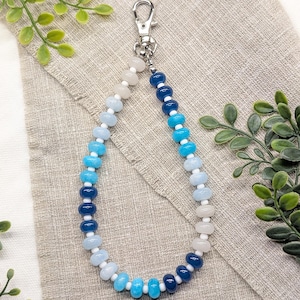 May include: A beaded lanyard featuring a silver-toned metal lobster clasp at the top. The lanyard is constructed from a repeating pattern of round, smooth beads in various shades of blue, white, and translucent grey, separated by small white spacers.