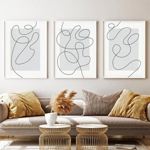 May include: Three minimalist line art prints in white frames on a white wall above a light grey sofa with brown throw pillows. The prints feature abstract black line drawings on a light grey background.