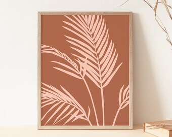 May include: A framed art print featuring a minimalist design of light pink palm leaves against a terracotta background. The artwork is encased in a light wood frame. A vase with dried grass and a book are on the table.