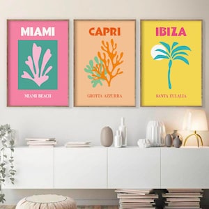 May include: Three framed prints with colorful illustrations of tropical plants and text. The first print is pink with a green background and says "Miami" and "Miami Beach". The second print is peach with a green and orange background and says "Capri" and "Grotta Azzurra". The third print is yellow with a blue background and says "Ibiza" and "Santa Eulalia".