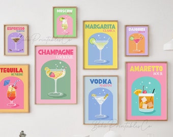 May include: Nine colourful printable posters featuring illustrations of different cocktails with their names in bold text. The posters are framed and displayed on a white wall above a wooden table.