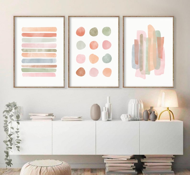 May include: Three abstract watercolor paintings in muted pink, peach, and green tones. The paintings are framed in natural wood and hung on a white wall above a white cabinet.