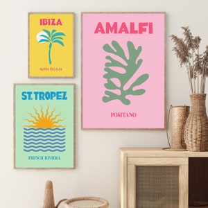 May include: Three framed prints with a summer vacation theme. The top print is pink with a green abstract leaf design and the text "AMALFI POSITANO". The middle print is yellow with a palm tree and the text "IBIZA SANTA EULALIA". The bottom print is green with a blue and yellow sun setting over the ocean and the text "ST. TROPEZ FRENCH RIVIERA".