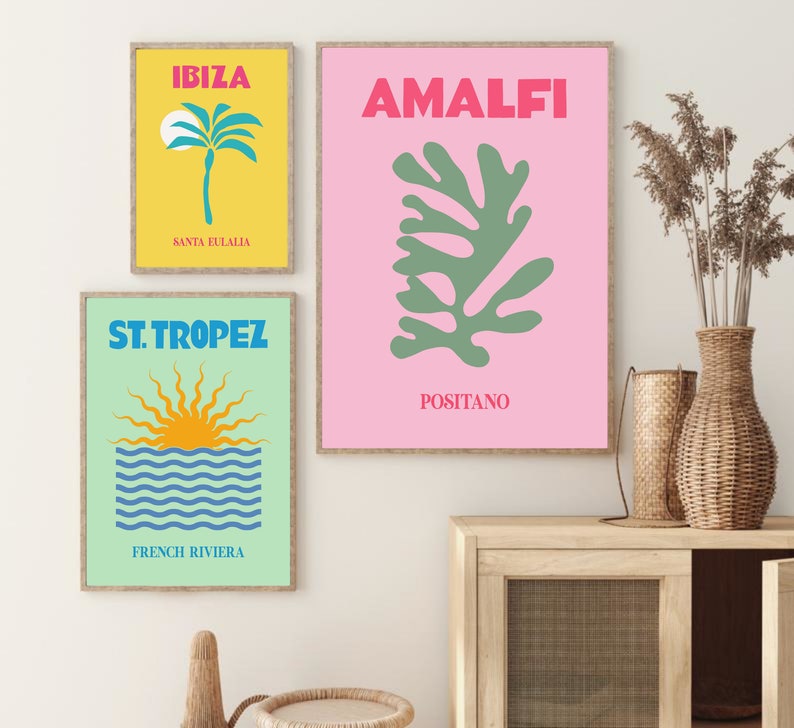May include: Three framed prints with a summer vacation theme. The top print is pink with a green abstract leaf design and the text "AMALFI POSITANO". The middle print is yellow with a palm tree and the text "IBIZA SANTA EULALIA". The bottom print is green with a blue and yellow sun setting over the ocean and the text "ST. TROPEZ FRENCH RIVIERA".
