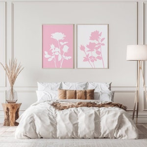 May include: Two framed prints of white rose silhouettes on a pink background. The prints are hanging on a white wall above a white bed with brown throw pillows and a brown blanket.