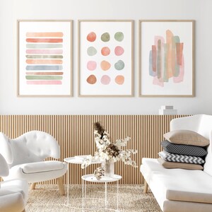 May include: Three framed abstract watercolor paintings with a neutral color palette of pink, green, and brown. The paintings are hung on a white wall above a wooden slat wall.
