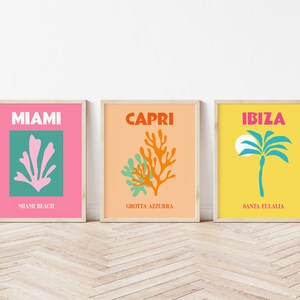 May include: Three framed prints with a minimalist design. The first print is pink with a green square in the center featuring a pink coral design. The text "MIAMI" is at the top of the print and "MIAMI BEACH" is at the bottom. The second print is peach with a green and orange coral design in the center. The text "CAPRI" is at the top of the print and "GROTTA AZZURRA" is at the bottom. The third print is yellow with a blue palm tree design in the center. The text "IBIZA" is at the top of the print and "SANTA EULALIA" is at the bottom.