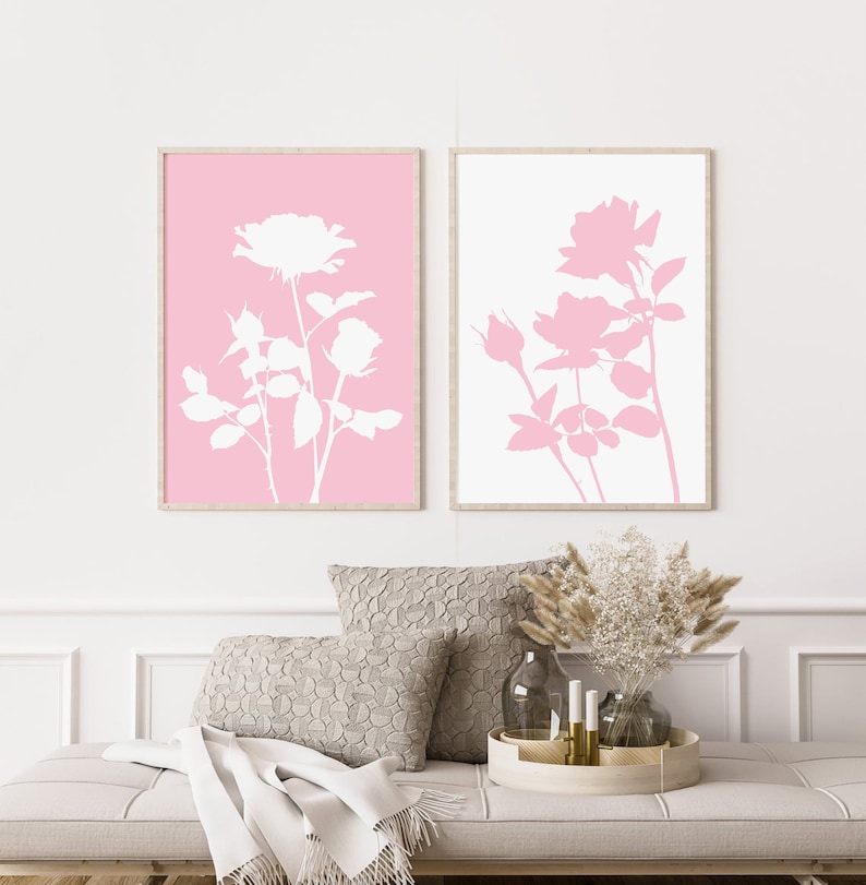 May include: Two framed prints with white silhouettes of roses on a pink and white background. The prints are hanging on a white wall above a couch with a throw blanket and pillows.