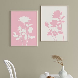 May include: Two framed prints of white rose silhouettes on a pink and white background. The prints are hanging on a light brown wall.