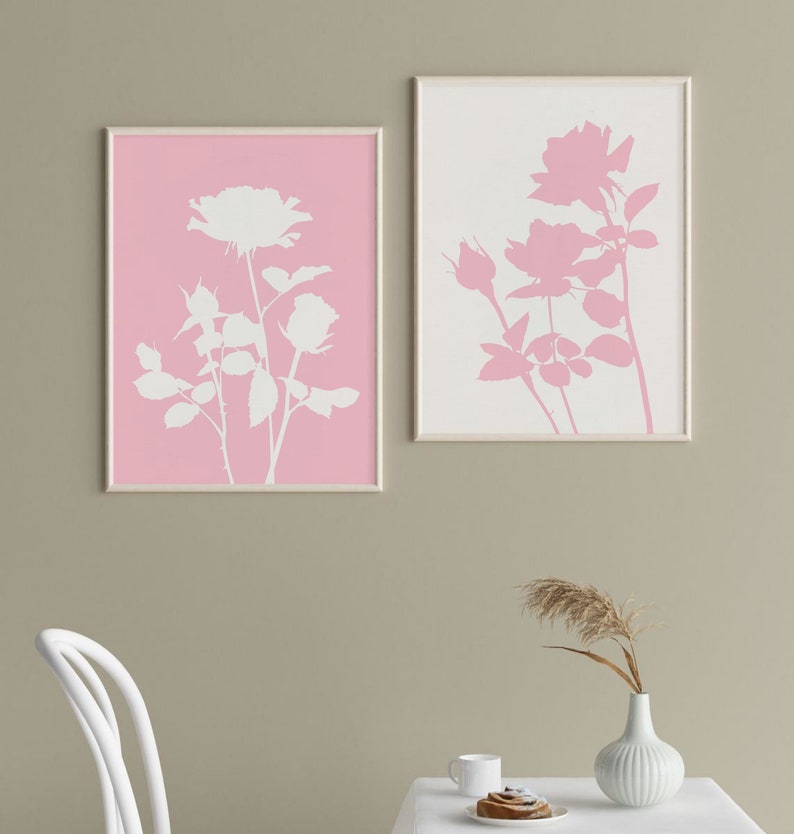 May include: Two framed prints of white rose silhouettes on a pink and white background. The prints are hanging on a light brown wall.
