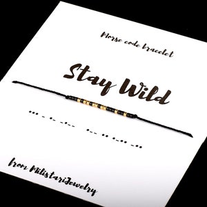May include: Black string bracelet with gold and black beads spelling out "Stay Wild" in Morse code. The bracelet is on a white card with the text "Morse code bracelet" and "from Militarijewelry".