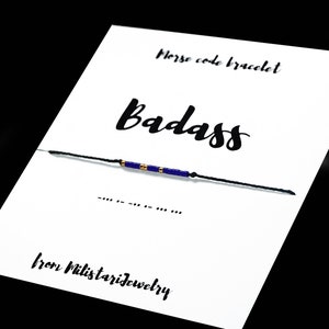 May include: A black string bracelet with blue and gold beads. The bracelet is on a white card with the word "Badass" written in black cursive and the text "Morse code bracelet" and "from Militarijewelry".