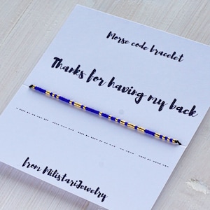 Peut inclure: Un bracelet d'amitié bleu et or avec le message "Merci d'avoir mon dos" en texte noir sur une carte blanche. La carte indique également "Bracelet en code morse" et "de Milistariejewelry".