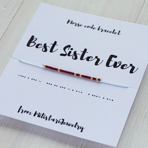 May include: A white card with the text "Morse code bracelet" and "Best Sister Ever" in black. A red and gold beaded bracelet is displayed on the card with the morse code for "Best Sister Ever" written below the bracelet. The card also includes the text "from Milistariejewelry".