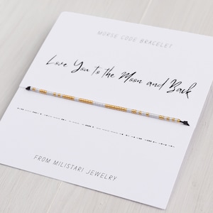 Peut inclure: Une carte blanche avec du texte noir qui dit "Bracelet en code Morse" et "Je t'aime jusqu'à la lune et retour". Un bracelet en perles blanches et dorées est présenté sur la carte. La carte indique également "De Miliatari Jewelry".