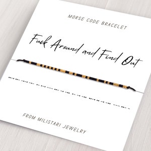 May include: A black and gold bracelet with the phrase "Fuck Around and Find Out" written in cursive on a white card. The bracelet is made of black and gold beads and is tied with a black string. The card also includes the phrase "Morse Code Bracelet" and "From Miliatari Jewelry".